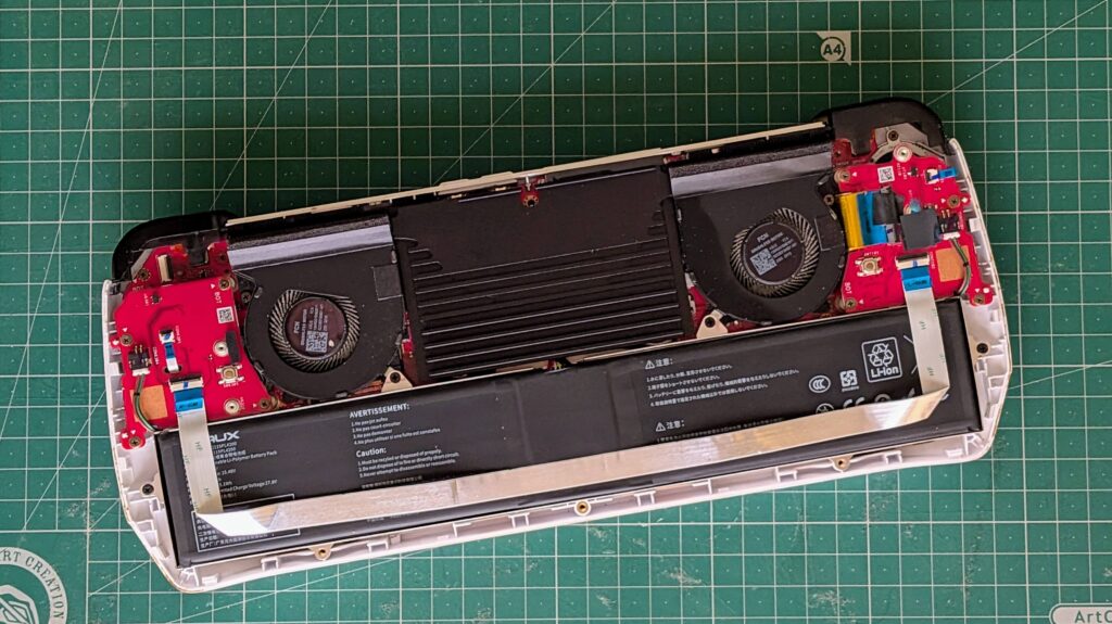 An opened electronic device showing internal components including fans, a battery, and circuit boards on a green cutting mat.