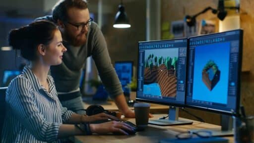 Two game developers working together at a dual-monitor setup, reviewing a 3D game scene in an editor.
