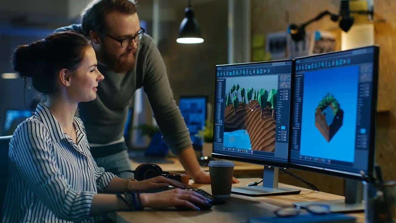 Two game developers working together at a dual-monitor setup, reviewing a 3D game scene in an editor.