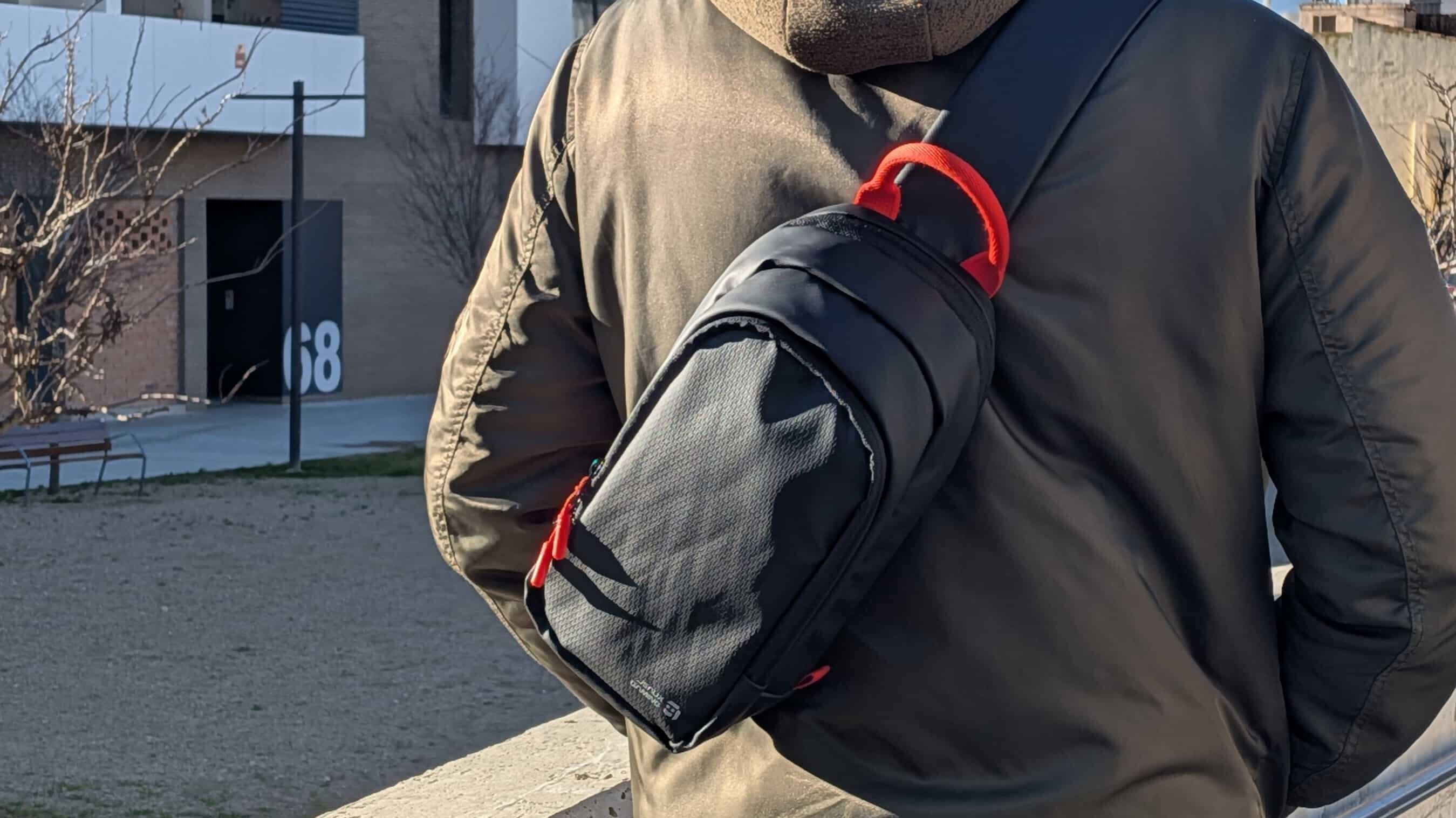 Man outdoors wearing a green jacket and brown hoodie with a black sling bag over his shoulder.