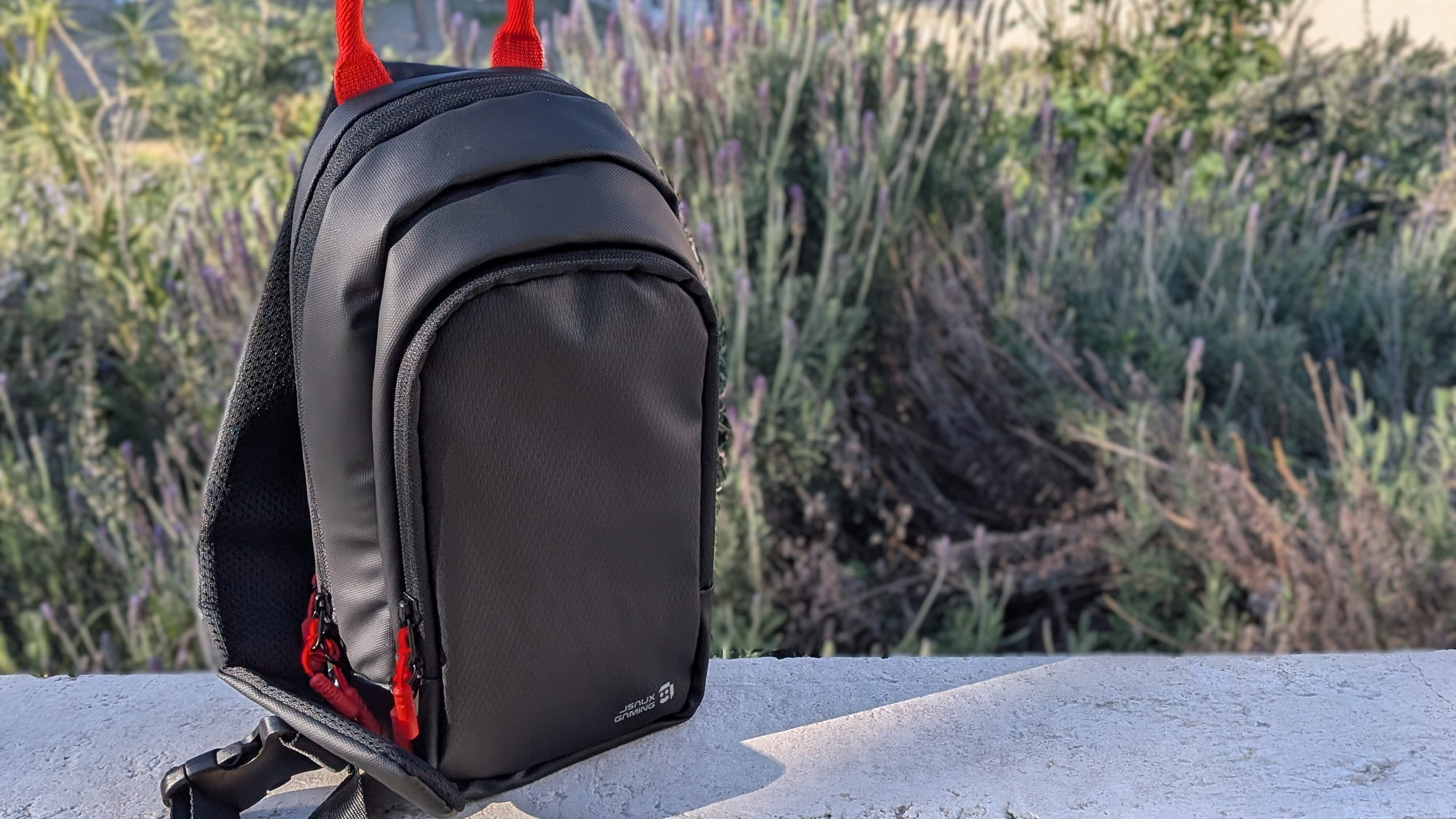 A small black backpack with red handles and zippers sits on a concrete ledge outdoors by green plants.