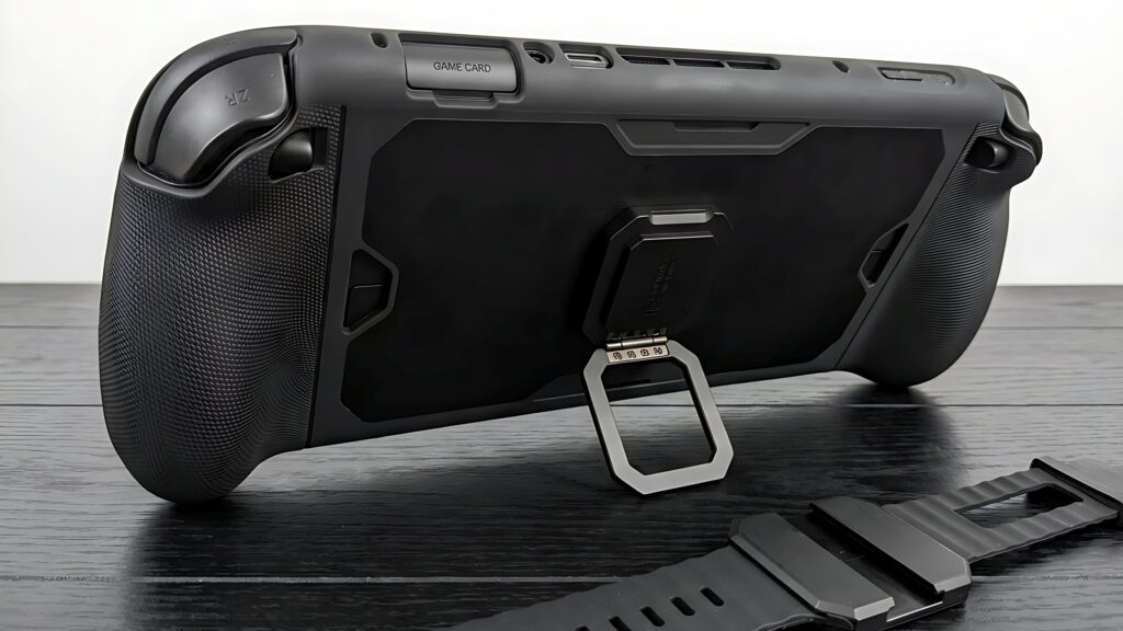 Close-up view of the back of a gaming handheld device with a kickstand and game card slot, placed on a wooden surface next to a black wrist strap.