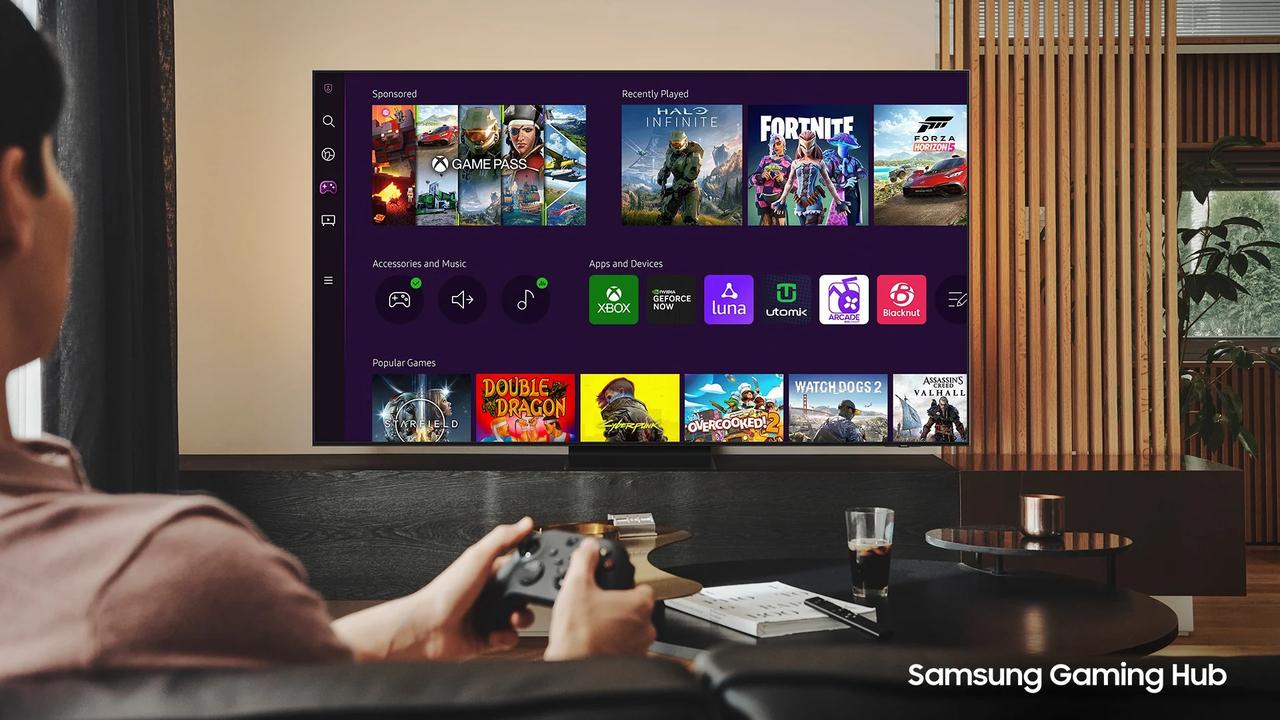A person sitting on a couch using a wireless controller to browse the Samsung Gaming Hub on a smart TV, featuring icons for Xbox, NVIDIA GeForce NOW, and Amazon Luna.