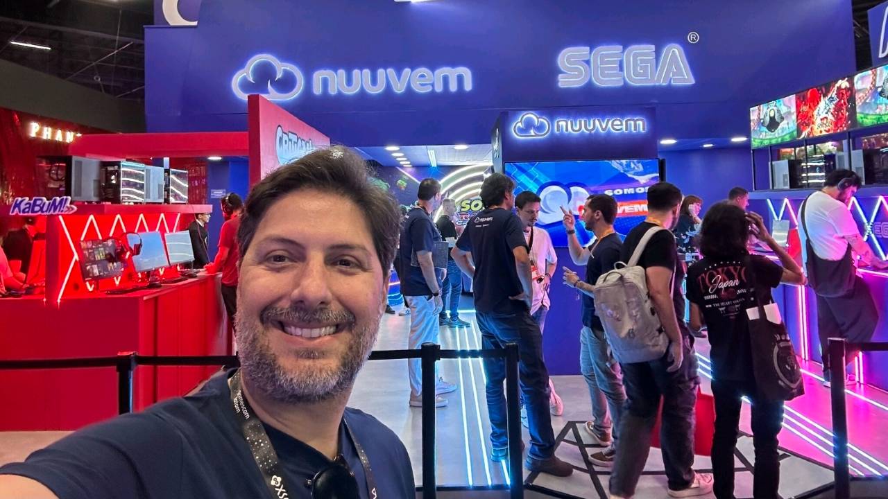 A selfie of Fernando Campos smiling man in a navy blue shirt standing in front of the glowing blue Nuuvem and SEGA signage. The background shows a busy convention floor with fans at gaming stations and the red CriticalLeap branding.