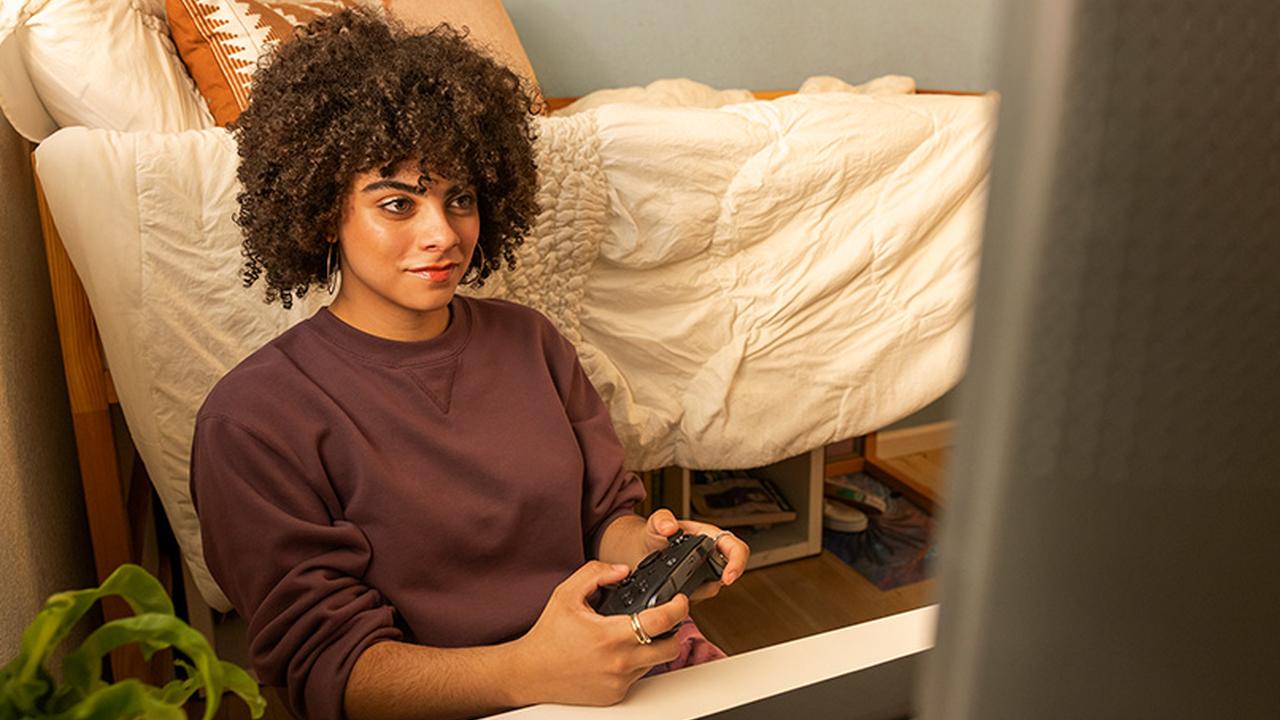 A woman holds a controller while playing XBOX on a screen in front of her.