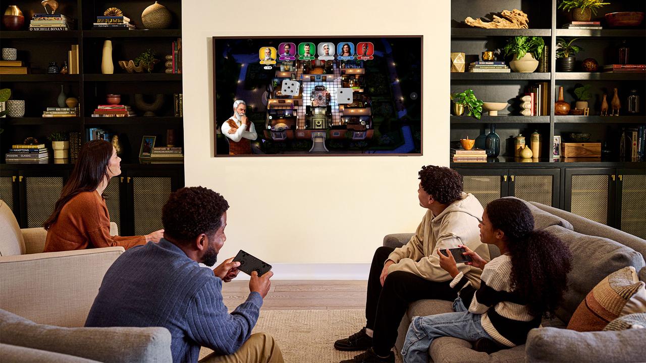Four people gathered in a living room playing a digital board game on a Samsung TV, with smartphones used as controllers.