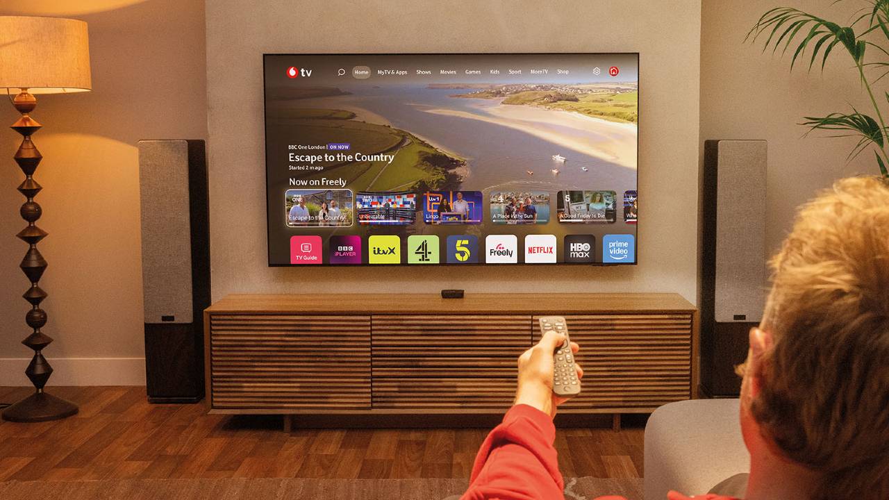 Man using a remote control with Vodafone TV displayed on a television in a living room.